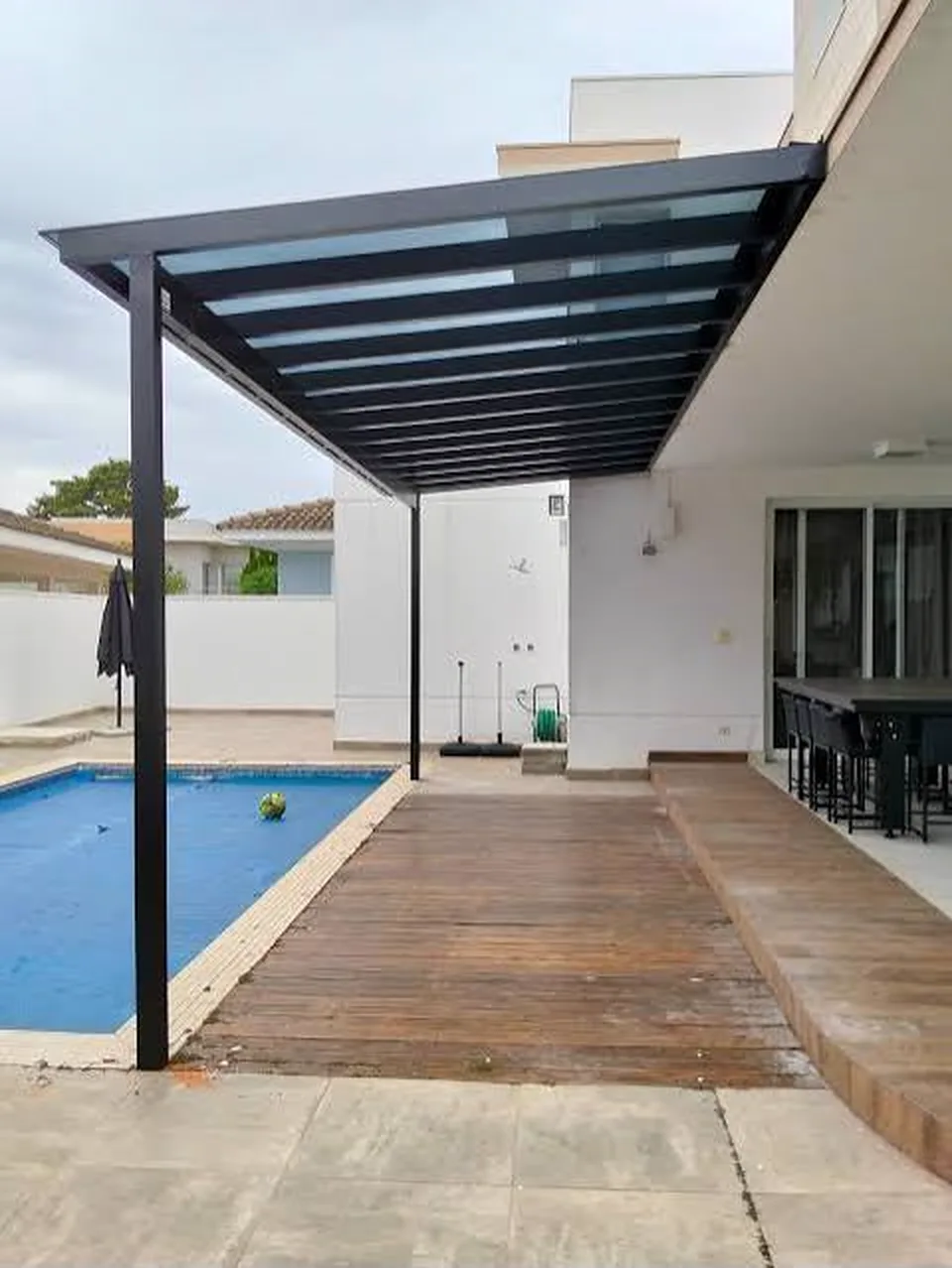 Cobertura de policarbonato com estrutura metálica escura sobre o deck da piscina, encostada na parede da casa